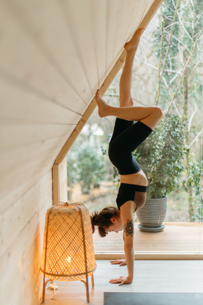 A person performs a handstand against a slanted wooden wall in a cozy room. They have one leg bent, wearing a black outfit. A lit wicker lamp is nearby, and a large potted plant sits in front of a window showing trees outside.