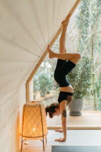 A person performs a wall-supported handstand in a cozy room with wooden walls and large windows. A lit wicker lamp sits on the floor nearby, and a potted plant is visible in the background.