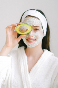 A person wearing a white robe and headband, with a creamy facial mask, holds an avocado half over one eye. They are smiling, and the background is plain and bright.