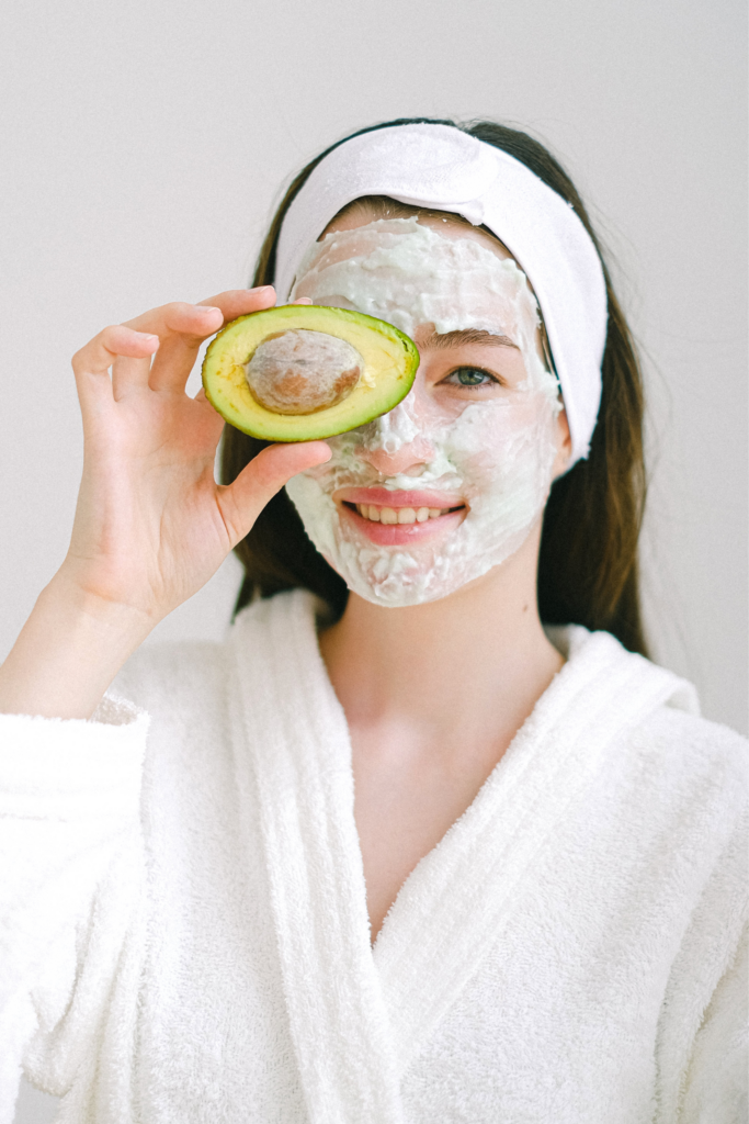 A person wearing a white robe and headband, with a creamy facial mask, holds an avocado half over one eye. They are smiling, and the background is plain and bright.