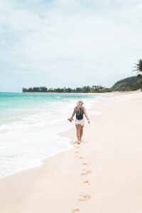 A person walks along a sandy beach, holding sandals and wearing a backpack. Footprints trail behind them in the sand as waves gently lap the shore. The scene is calm, with a clear sky and distant trees along the coastline.