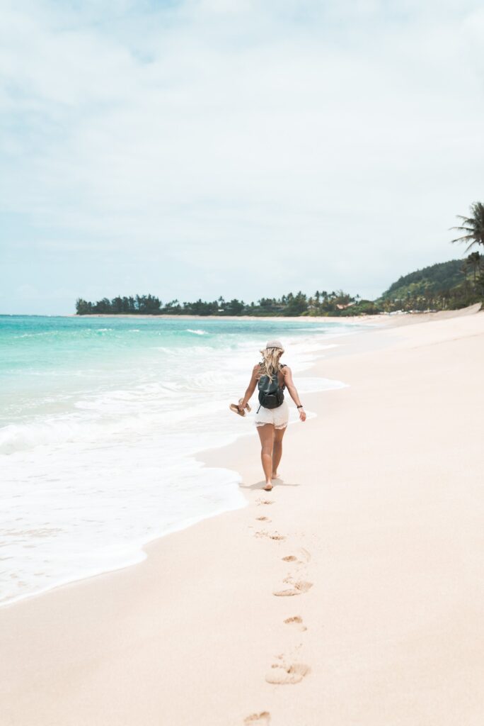 A person walks along a sandy beach, holding sandals and wearing a backpack. Footprints trail behind them in the sand as waves gently lap the shore. The scene is calm, with a clear sky and distant trees along the coastline.