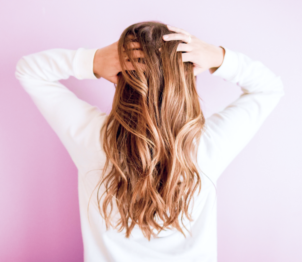 A person with long, wavy brown hair stands against a pastel purple background, facing away. Their white top highlights their flawless skin as they playfully run their hands through their hair.