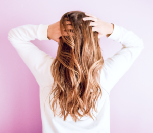 A person with long, wavy brown hair stands against a pastel purple background, facing away. Their white top highlights their flawless skin as they playfully run their hands through their hair.