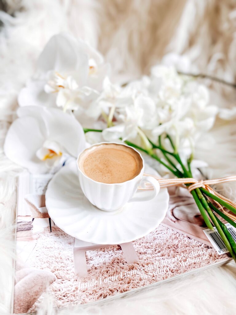 A white cup of coffee sits on a saucer, placed on an open magazine. Beside it are white orchids, set against a soft, fluffy background, creating a cozy and elegant atmosphere.