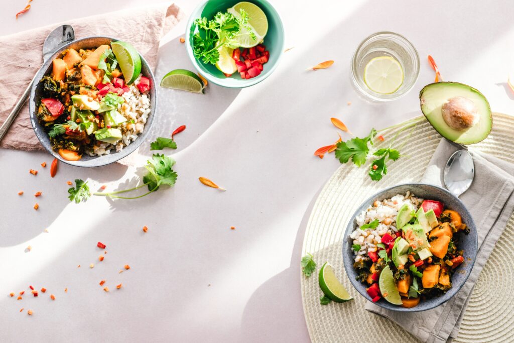 Two bowls of vibrant vegetable salad with avocado slices, lime wedges, and cilantro on a light surface. A cut avocado, a bowl with lime and diced tomatoes, and a glass with lemon water are nearby. Scattered ingredients add to the fresh, colorful presentation.
