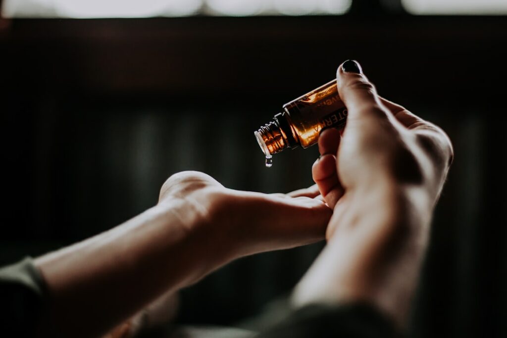 A person holding a small, brown glass bottle and pouring a few drops of essential oil into their palm. The background is softly blurred, highlighting the focus on the hands and the bottle.