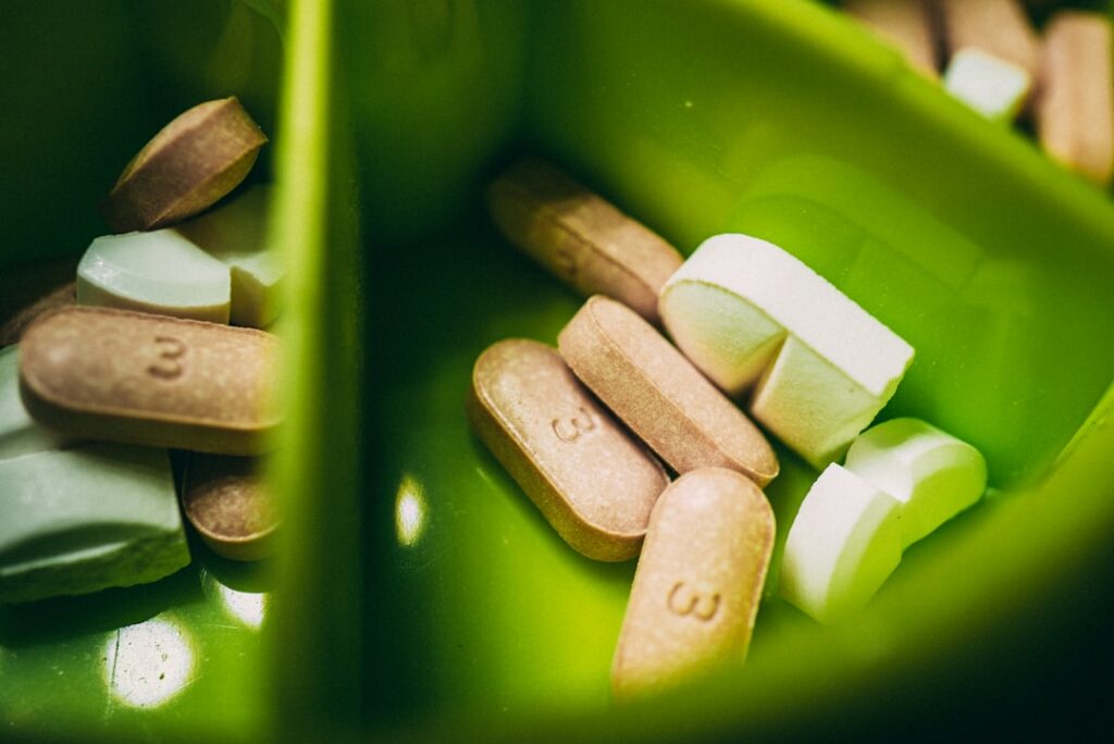 A close-up of various pills and tablets in a green divided container. The pills are of different shapes and colors, including white oblong and beige oval ones, some marked with the number "3.