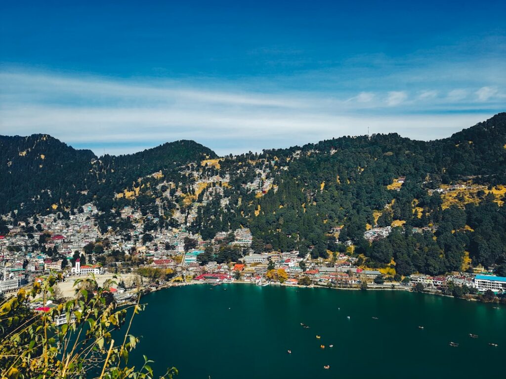 Scenic view of Nainital, featuring a tranquil lake surrounded by lush green hills and colorful buildings. Small boats float on the water under a clear blue sky, creating a peaceful and vibrant atmosphere.