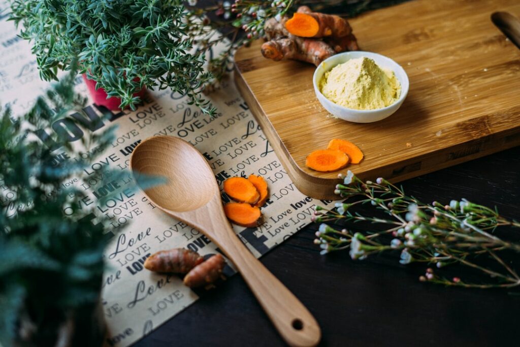 A wooden spoon and turmeric roots with sliced pieces are on a table. A small bowl of turmeric powder is placed on a wooden cutting board. Surrounding the items are sprigs of herbs and a "LOVE" patterned cloth in the background.