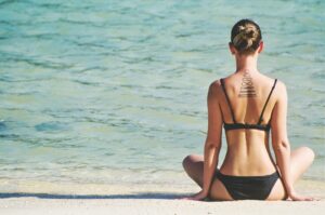 A woman in a black bikini sits on a sandy beach, facing the turquoise ocean. Her hair is tied up, and she has a geometric tattoo on her upper back. The scene is serene and sunlit.