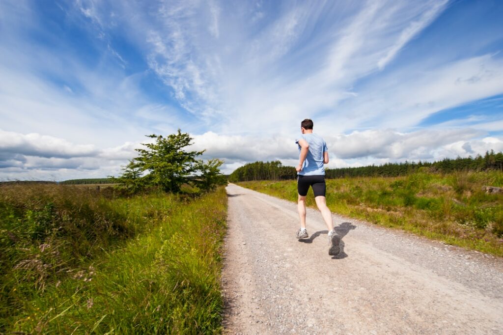 A person is jogging away on a gravel path surrounded by green fields and trees. The sky is vast and filled with scattered clouds. The scene conveys a sense of openness and freedom.
