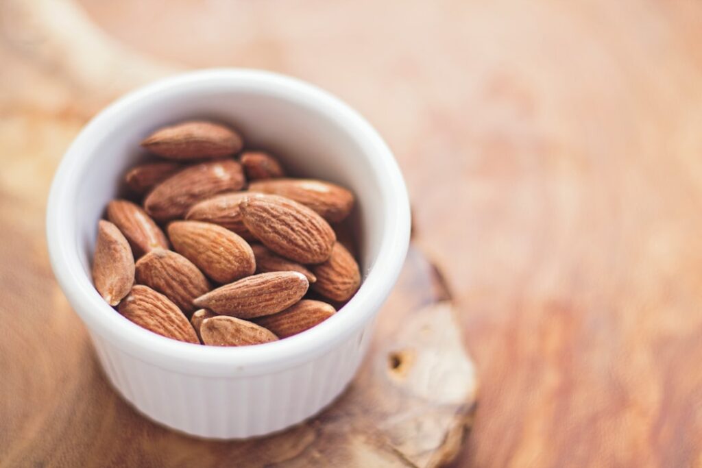 A small white bowl filled with whole almonds sits on a wooden surface. The almonds are brown and textured, contrasting with the smooth, white bowl. The background is softly blurred, highlighting the nuts.