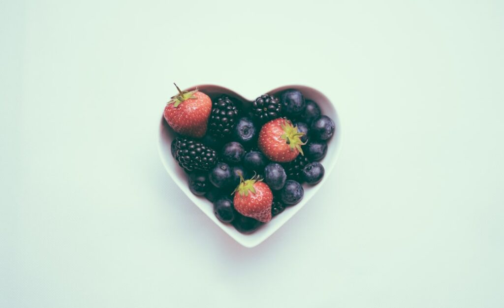 A heart-shaped bowl filled with fresh strawberries, blueberries, and blackberries against a light background.