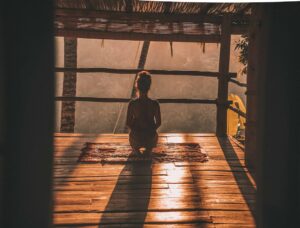 A person sits cross-legged on a woven mat, facing a sunrise. The scene is set in a wooden structure with a thatched roof. Warm sunlight filters through, casting long shadows and illuminating surrounding trees.