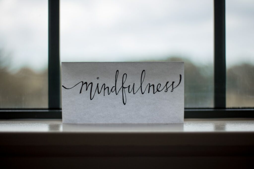 A card with the word "mindfulness" written in elegant script is placed on a window sill. The background shows a blurred view of the outdoors, suggesting a calm and serene atmosphere.