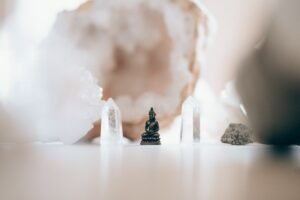 A small Buddha statue is surrounded by translucent crystals and stones on a light surface. The background is softly blurred, creating a serene and peaceful atmosphere.