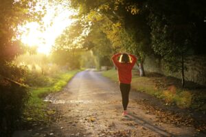 A person in a red sweatshirt and black pants walks down a tree-lined path during sunset. Golden light filters through the leaves, illuminating fallen leaves on the path. The scene conveys a peaceful, autumnal atmosphere.