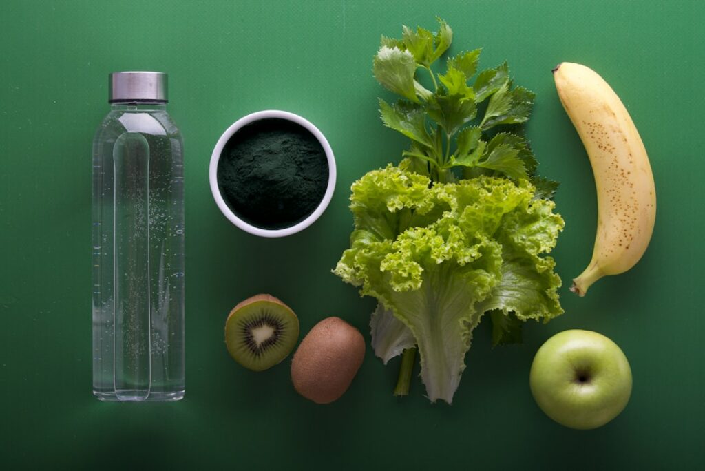 A flat lay of healthy items on a green background includes a water bottle, a bowl of green powder, leafy greens, a banana, a kiwi, an apple, and a brown egg.