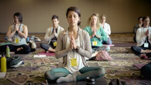 A group of people sits cross-legged on yoga mats in a conference room, meditating with eyes closed and hands in prayer position. They appear relaxed and focused, surrounded by personal items like water bottles and bags.