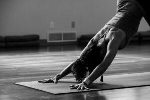 Black and white image of a person practicing yoga in a studio. They are on a mat in the downward dog position, with their hands and feet on the ground, creating an inverted V shape with their body. The room has wooden flooring.
