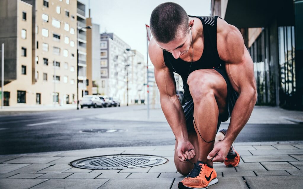 A person in athletic wear kneels on a city sidewalk, tying their orange running shoe. They're wearing headphones, and buildings and parked cars are visible in the background.